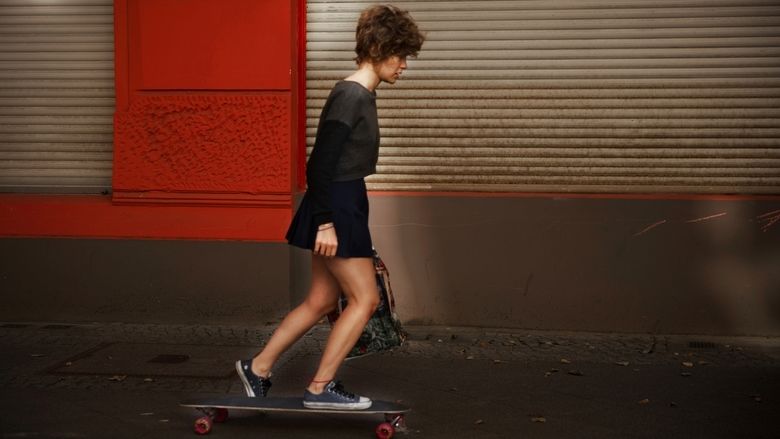 Carla Juri riding on a skateboard while wearing a black long sleeve shirt and black skirt in a scene from the 2013 film, Wetlands