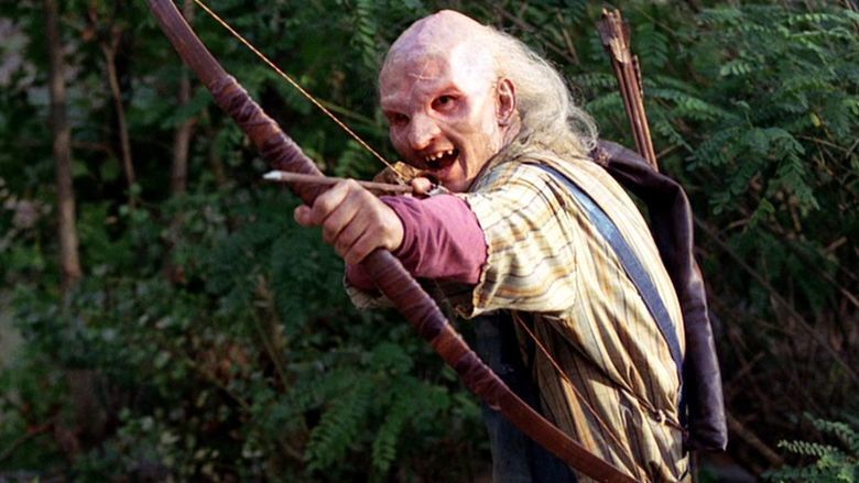 In the movie scene of Wrong turn 6 last Resort 2014, three finger (left) is happy smiling, holding a bow and arrow attempting to shoot with a quiver in his back in the middle of the forest, has white hair deformed and old scar face wearing a maroon long sleeve under a yellow striped long sleeve polo and a jumper.