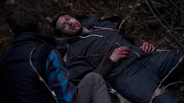 In the movie scene of Wrong turn 6 last Resort 2014, on the left a man crouching and looking beside Anthony Ilott has black hair wearing winter sleeveless jacket over blue checkered long sleeve polo, on the right Anthony Ilott lying on the ground with blood on his hand and face with barbed wired over them, has brown hair wearing black hoodie and denim pants.