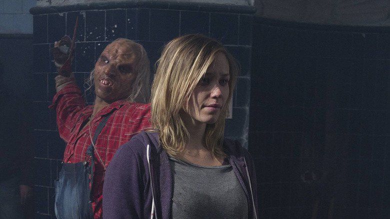 In the movie scene of Wrong turn 6 last Resort 2014, three finger (left) is behind Aqueela Zoll (right) holding pickaxe attempting to hit her behind has white hair deformed face wearing blue jumper and a red checkered long sleeve, Aqueela Zoll (right) is scared while standing has brown hair wearing gray shirt and purple jacket.