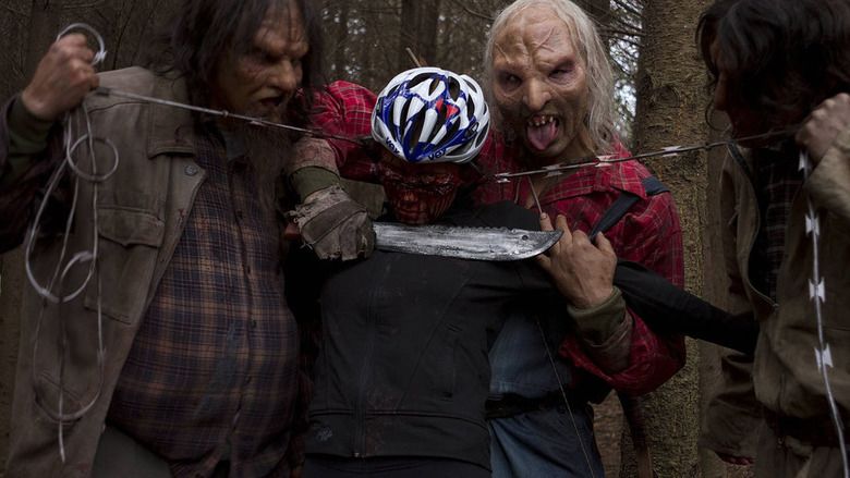 In the movie scene of Wrong turn 6 last Resort 2014, from the left is One eye (1st) is standing holding the barbed wire with his right hand has shoulder length black hair, wearing a black ragged jacket, next to him is a dead woman (2nd) has barbed wire wrapped around his head wear a cycling helmet and a long sleeve cycling shirt, three finger (3rd) is excited with his tongue out while holding a machete with his right hand and hold the left hand of the dead man with his left hand, has white long hair deformed face with old scars on it wearing jumpers and red checkered long sleeve, saw tooth (4th) is standing looking at the dead man while holding the barbed wire with his left hand has long black hair wearing a checkered shirt and a brown jacket.