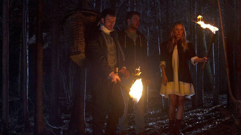In the movie scene of Wrong turn 6 last Resort 2014, on the left is Chris Jarvis is looking and holding the torch down with a big basket on his back, has brown hair wearing gray shirt polo black pants and black jacket, in the middle is Anthony Ilott standing while looking at Sadie Katz (right) has brown hair wearing polo shirt and dark blue hoodie jacket, on the right is Sadie Katz standing while holding the torch with her left hand has long blond hair wearing a white dress, pair of boots and black jacket.