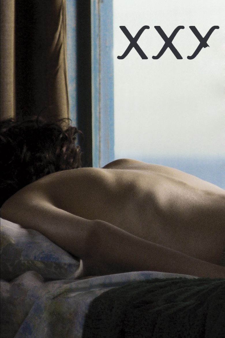 Ines Efron as Alex Kraken having a curly hair, lying on the bed in a back position with a brown curtain and window in the background, the word "XXY" written on the right side, and Ines is topless