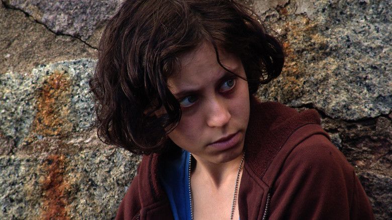 Ines Efron as Alex Kraken looking at someone with a serious face having a short curly hair and a rock in the background, wearing a blue shirt under a brown jacket, and a silver necklace