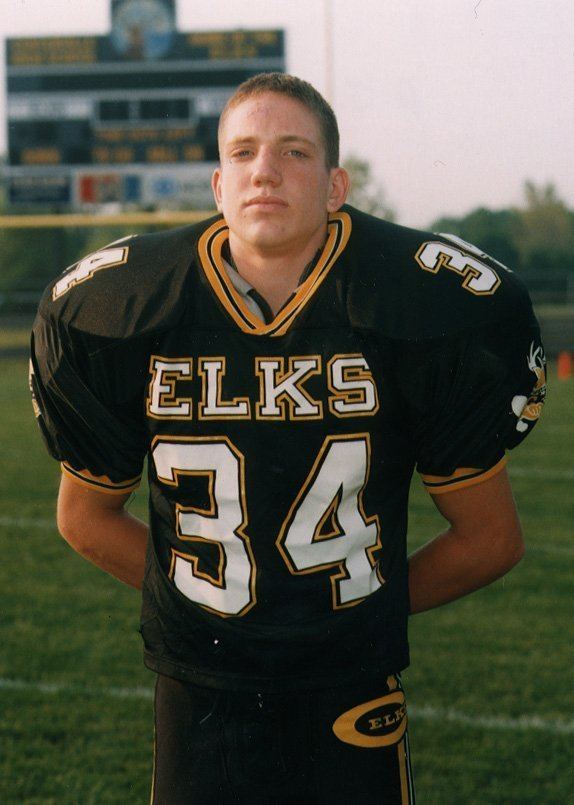 Young A J Hawk seriously looking and standing while both arms behind his back, has blonde hair wearing pads under a black jersey with an “ELKS 34” on it so as his black pants