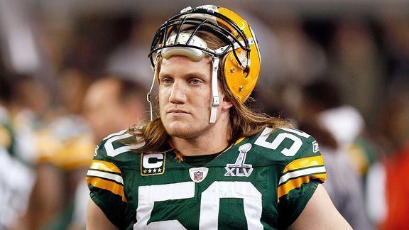 A J Hawk seriously looking, has short blonde hair wearing a yellow helmet, a black shirt, and pads under a green jersey with a #50 on it