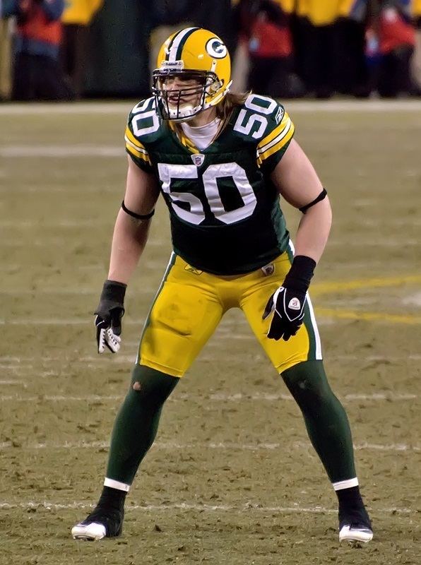 A J Hawk standing with wide-open legs inside the football field, he has short blonde hair wearing a yellow helmet, a white shirt and pads under a green jersey with a #50 on it, black gloves, a black-tie on both arms, thigh pads under a yellow fitted short, a green long socks and a black cleats