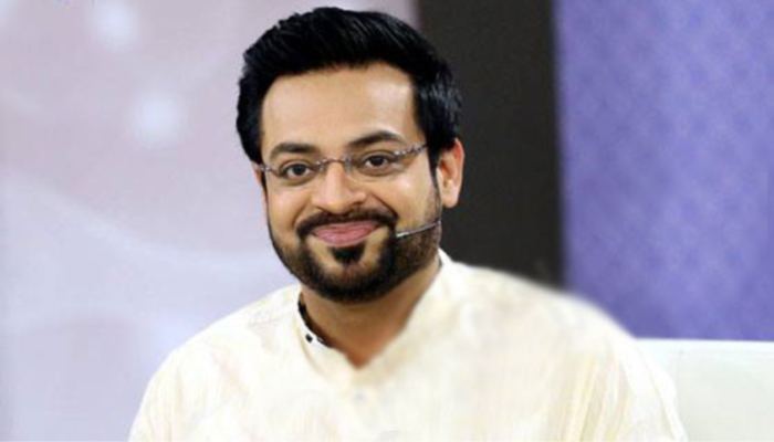 Aamir Liaquat is smiling, has black hair, a beard, and a mustache, uses a wireless microphone, wearing eyeglasses and white shalwar kameez.