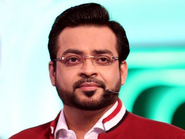 Aamir Liaquat is serious, has black hair, a beard, and a mustache, uses a wireless microphone, and wears eyeglasses, and white long sleeves under a red jacket.