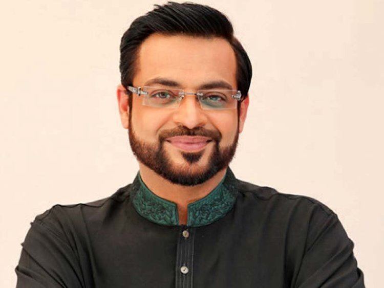 Aamir Liaquat is smiling, has black hair, a beard, and a mustache, wearing eyeglasses, black shalwar kameez with a green collar design.