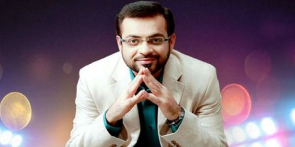 Aamir Liaquat is smiling, has black hair, a beard, and a mustache, wearing eyeglasses, a wristwatch on his left hand, and green long sleeves under a white suit.