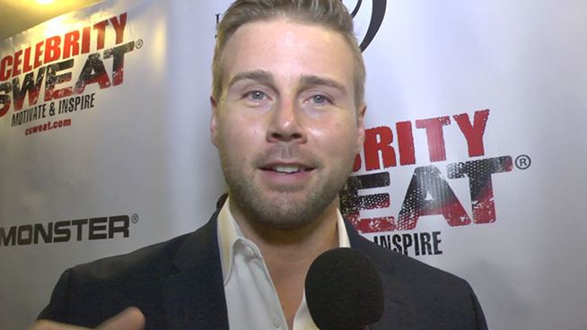 Aaron Hill with short blonde hair, wearing a black suit and a white shirt with a microphone in front of him.