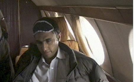 Abdolmalek Rigi inside an airplane on the way to Iran wearing a brown jacket over a white shirt and an eye cover.