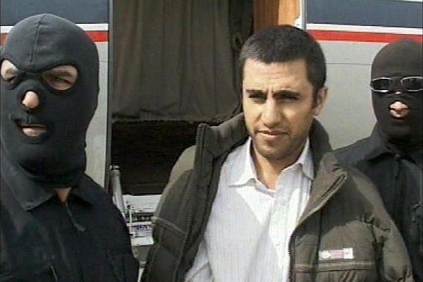 Abdolmalek Rigi being escorted by military personnel in Iran on February 23, 2010 wearing a brown jacket over a white shirt.