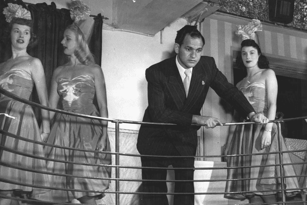 Abe Saffron leaning on the railing while three women walking behind him and he is wearing a black coat, long sleeves, necktie, and pants