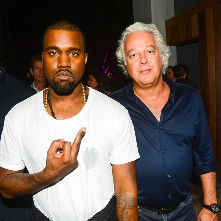 Kanye West with a serious face while showing a middle finger while Aby Rosen with a tight-lipped smile and with people at the back. Kanye with beard and mustache, a tattoo on his arm, wearing necklaces, a white shirt, and black pants while Aby with white hair, wearing a watch, blue folded long sleeves, a black belt, and blue jeans.