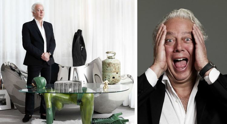 On the left, Aby Rosen is standing while his hands holding each together beside a couch and a table, with a serious face, with white hair, wearing a watch, a black coat over white long sleeves, black pants, and black shoes.On the right, Aby Rosen with a surprised face while his hands on it, with white hair, wearing a watch, and a black coat over white long sleeves.