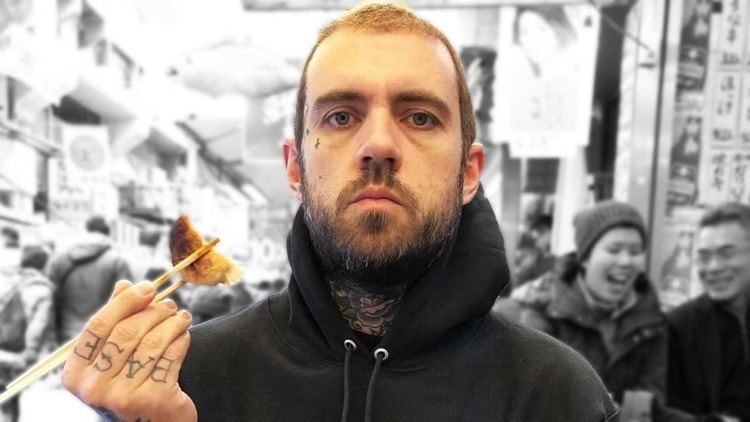 Adam Grandmaison is serious. He is holding chopsticks with dumplings, has a mustache and beard, a tattoo on his neck and fingers, and is wearing a black jacket.
