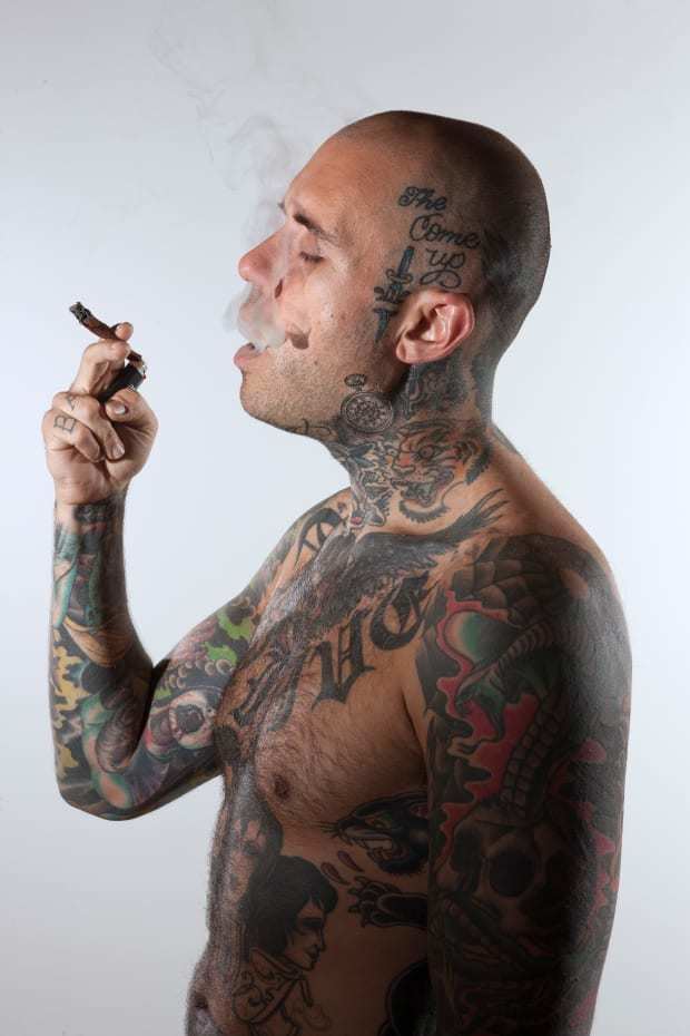 Adam Grandmaison is serious, side view, with closed eyes while smoking, has bald head and with tattoo all over his body top naked.