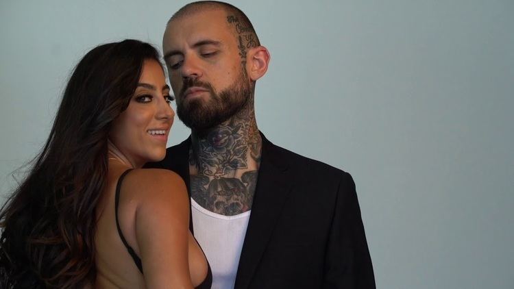 On a gray background, on the left, Lena Nersesian is smiling. Standing in her back view with her side breast visible, looking to her right, she has long black hair and is wearing a black noodle strap top and a gold necklace. On the right, Adam Grandmaison is standing, looking at Lena, he has a bald head, a beard, and a mustache, he has a tattoo from his neck to his head, and he is wearing a tank top under a black coat.