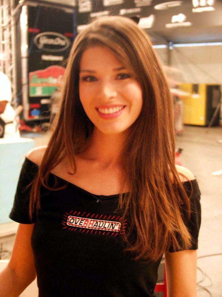 Adrienne Janic smiling with a golden-brown straight hair while wearing a black off-shoulder blouse with an overhaulin' print on the center