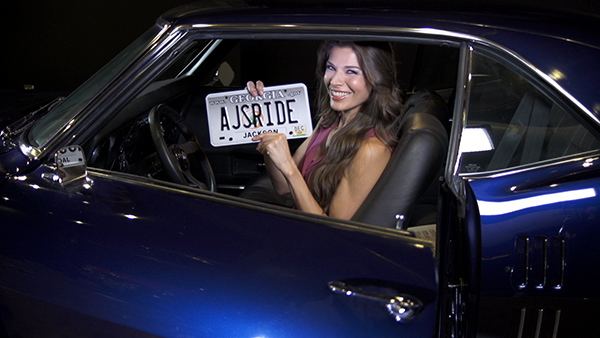 Adrienne Janic smiling and sitting on the driver's seat while holding a plate number "AJSRIDE", with black wavy hair, and wearing a purple blouse