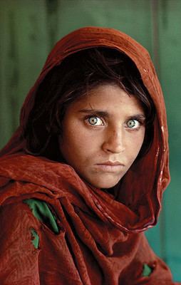Afghan Girl with green eyes staring directly into the camera, and wearing a red scarf draped loosely over her head, and green shirt.