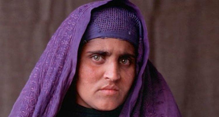 Sharbat Gula also known as the "Afghan Girl" with a serious face, green eyes, and wearing a purple scarf and black shirt.