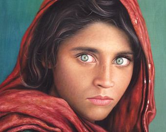 Afghan Girl with green eyes staring directly into the camera, and wearing a red scarf draped loosely over her head.