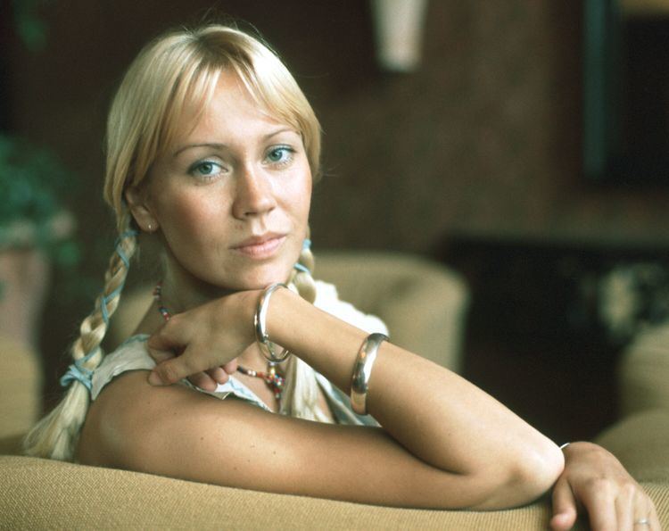 Agnetha Faltskog sitting on the couch while hand on her chin and wearing a white sleeveless blouse, beaded necklace, and bracelets