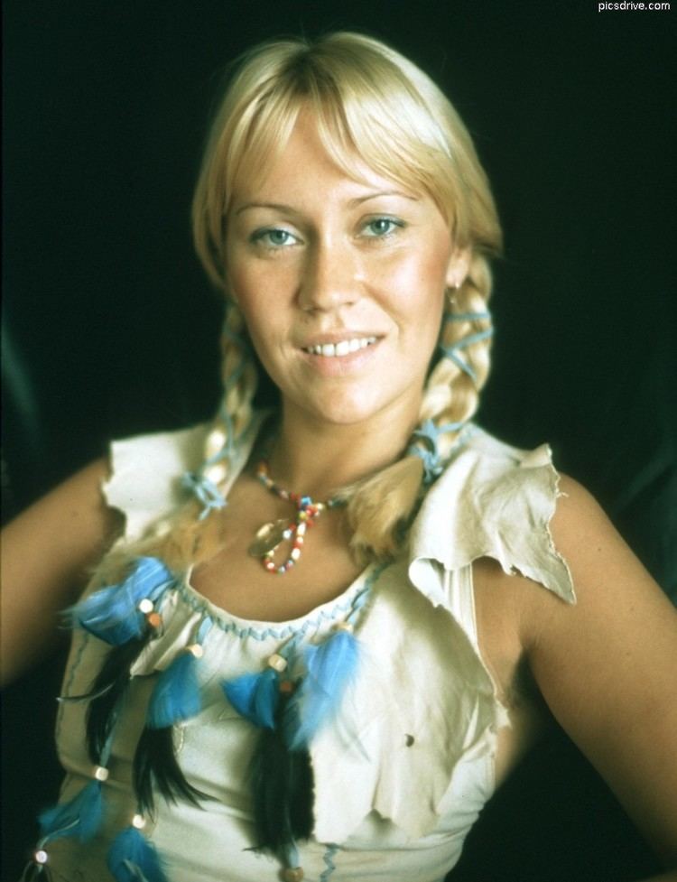 Agnetha Faltskog smiling with braided hair while wearing a white sleeveless blouse with black and blue feathers and beaded necklace