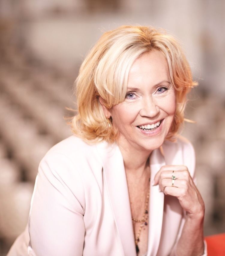 Agnetha Faltskog smiling while wearing a pink coat, necklace, earrings, and ring