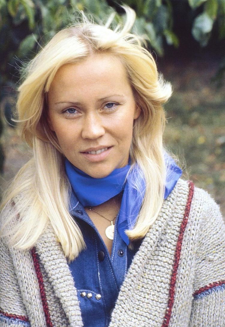 Agnetha Faltskog smiling while wearing a gray and maroon knitted blazer, denim blouse, blue scarf, and necklace