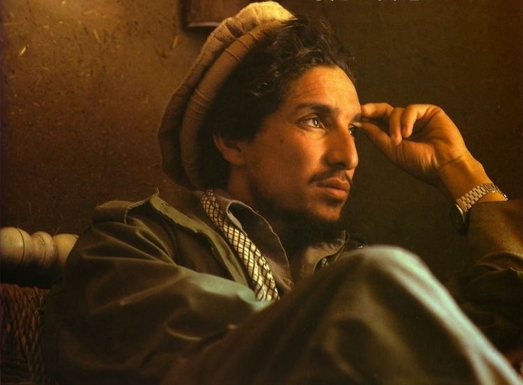 Ahmad Shah Massoud inside a building wearing a brown desert hat along with a striped sloth and green jacket and a watch.