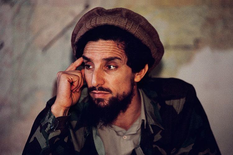 Ahmad Shah Massoud wearing a brown desert hat and an army jacket with thicker facial hair and his finger in his head.