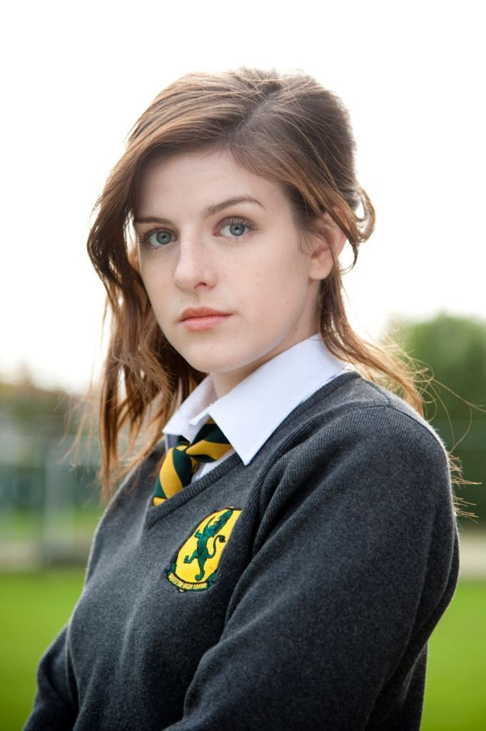 Aisling Loftus is serious, has brown hair, wearing her uniform, a white polo, green and yellow necktie under a gray jacket with a lion print on left.