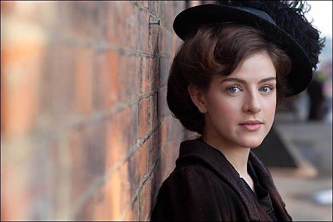 Aisling Loftus is serious, has brown hair, and wearing a black hat, and black top under a brown suit.