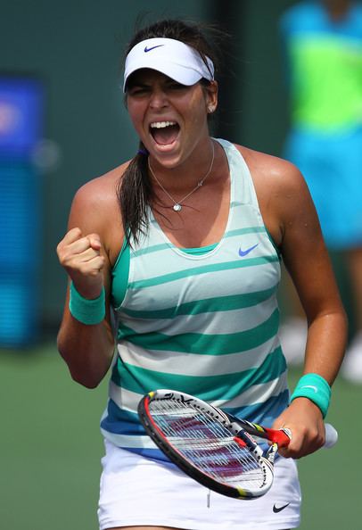 Ajla Tomljanovic shouting while holding a racket and wearing a white and green top, white cap, and white skirt