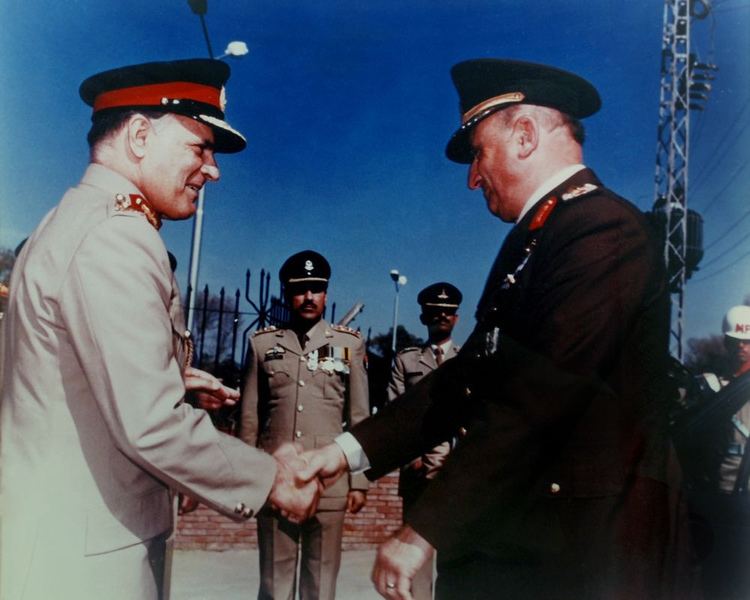 Akhtar Abdur Rahman smiling while shaking hands with the Turkish Defense Chief in 1988 with three soldiers standing straight in the background wearing black peaked caps and brown coats with badges. Akhtar is wearing a black peaked cap and brown long sleeve with insignia and badges while the Turkish Defense Chief is wearing a black peaked cap and white long sleeve under a black coat with insignia
