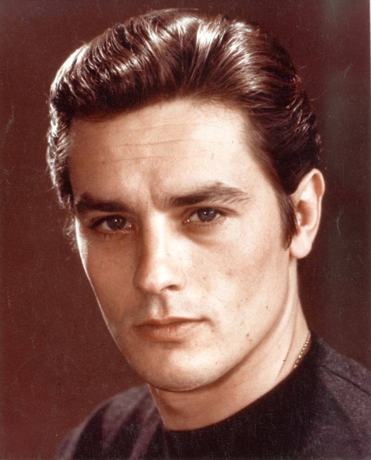 Young Alain Delon with a serious face, wearing a gold necklace and a black shirt.