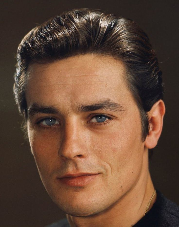 Young Alain Delon smiling, with blue eyes, wearing a gold necklace and a black shirt.
