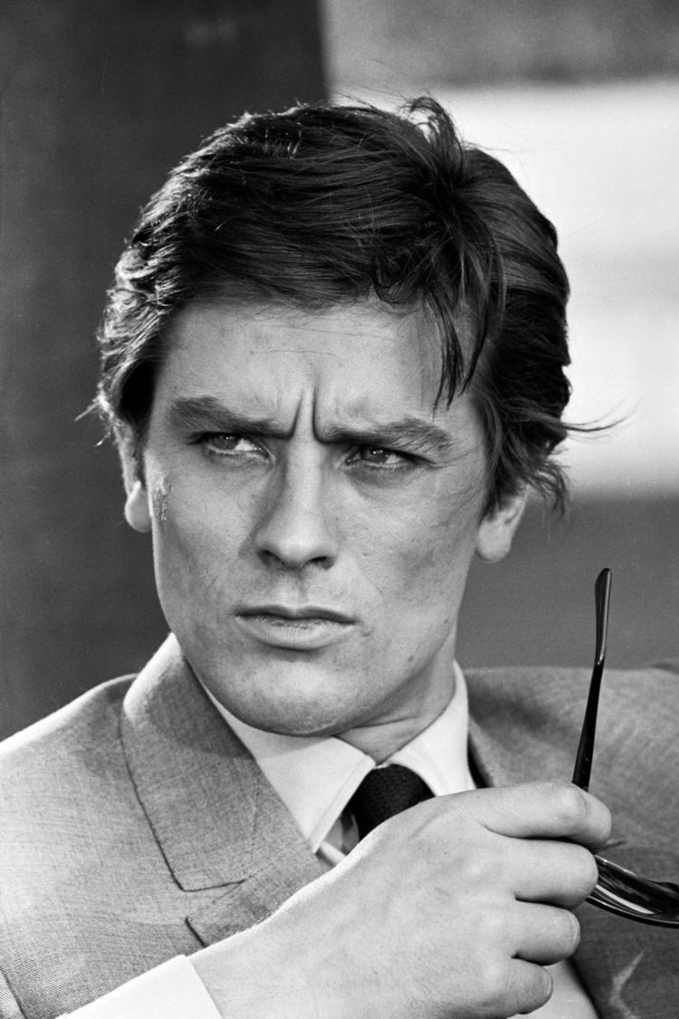 Alain Delon with a grumpy face while holding sunglasses, wearing a coat over white long sleeves and a black tie.