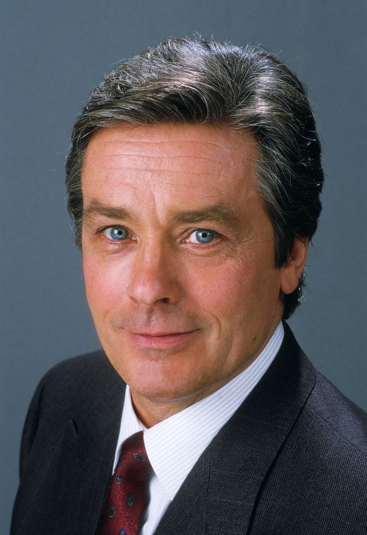 Young Alain Delon smiling, with blue eyes, wearing a black coat over white long sleeves and a red tie.