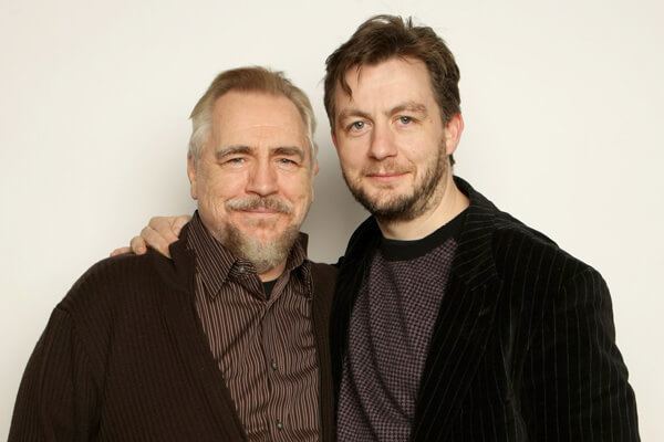 Brian Cox and Alan Cox are smiling with mustaches and beards. Brian is wearing a brown striped long sleeve under a dark brown cardigan while Alan is wearing a black checkered shirt under a black striped coat