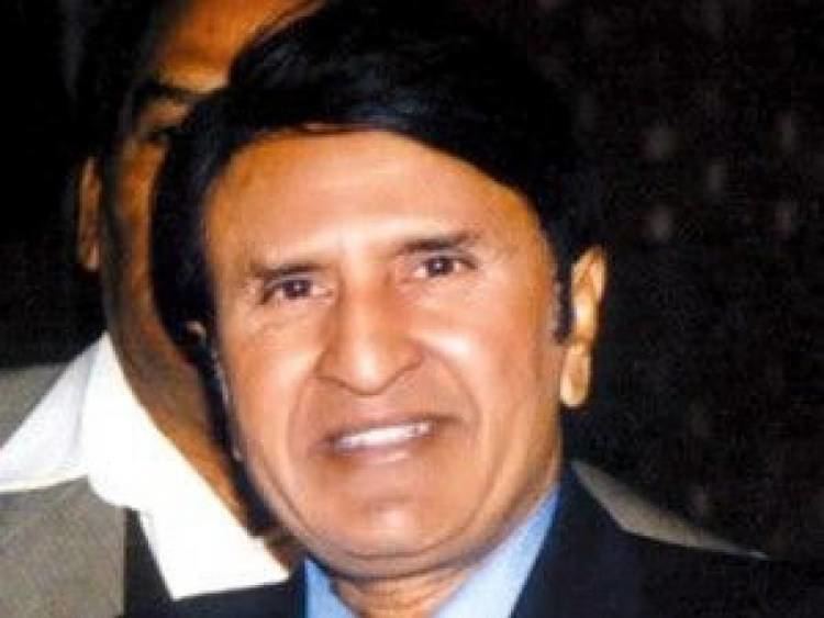 Albela smiling and wearing a blue shirt under a black coat