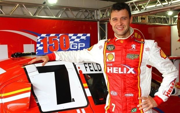 Alceu Feldmann smiling while leaning on the car and wearing a white, red, and yellow jacket