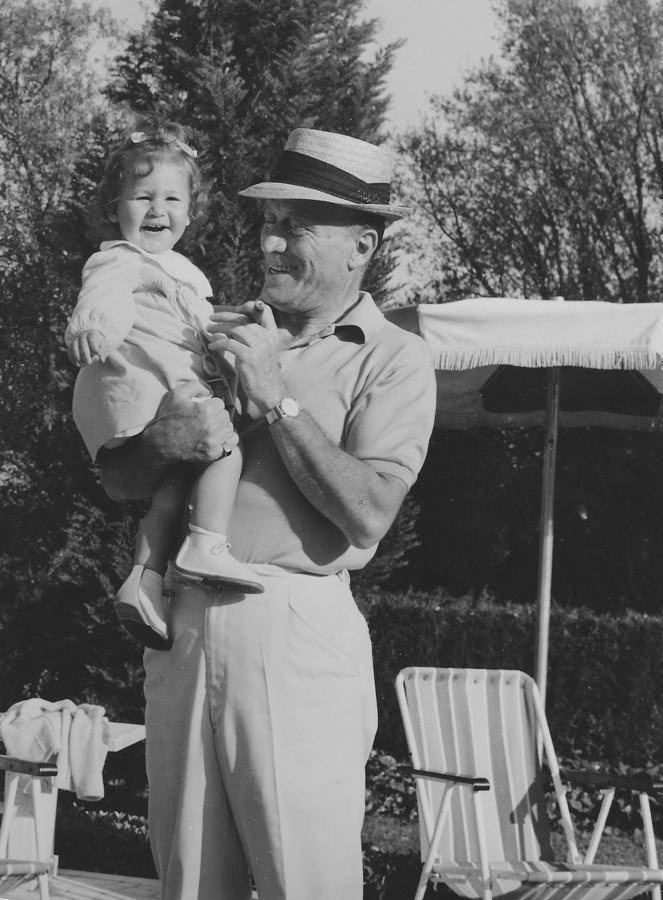 Aldo Gucci smiling while carrying at his daughter Patricia Gucci with a giggling face. Aldo is wearing a hat, watch, polo shirt, and white pants while Patricia is wearing a dress and white shoes.