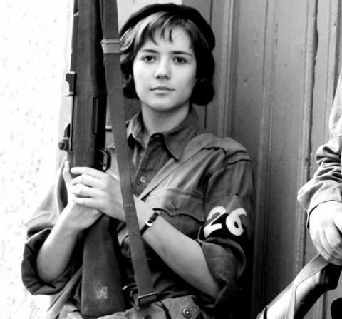 Aleida March with a serious face while holding a riffle, wearing a black hat, and an army uniform.