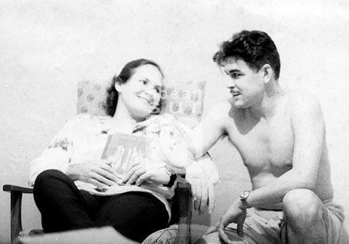 Aleida March and Che Guevara are looking at each other. Aleida sitting on a chair, holding a book, wearing a white long sleeve blouse and black pants while Che is topless.