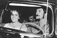 Aleida March smiling while Che Guevarra smoking with a man inside a car. Aleida wearing a pearl necklace and a white dress while Che with a beard and wearing a long sleeve.